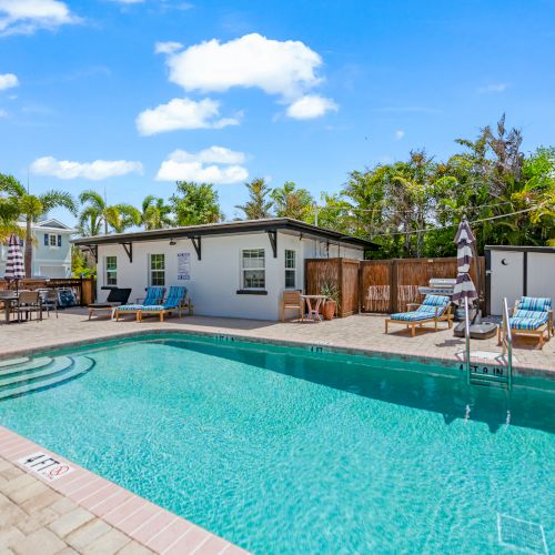 A backyard pool with lounge chairs, umbrellas, and palm trees under a blue sky with clouds.