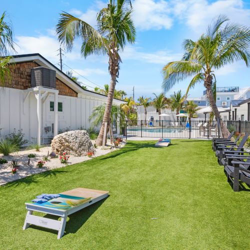 A grassy area with palm trees, cornhole game, and Adirondack chairs, leading to a pool with a fence around it under a clear blue sky.