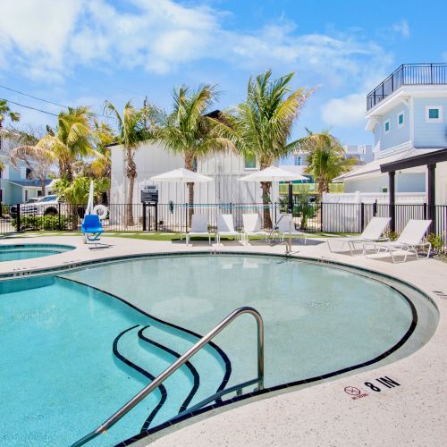 Outdoor pool area with lounge chairs, palm trees, and nearby blue buildings under a clear sky. Peaceful resort-like setting.