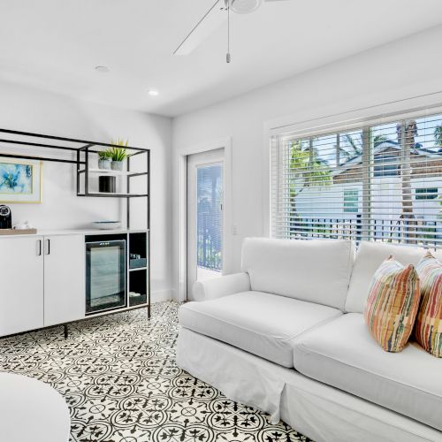 A bright room with a white sofa, decorative pillows, patterned floor, cabinet, refrigerator, and wall art. Large window with blinds.