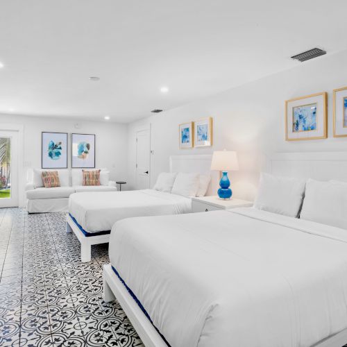 The image shows a bright hotel room with two beds, patterned floor tiles, a sofa, artwork on the walls, and a small dresser.