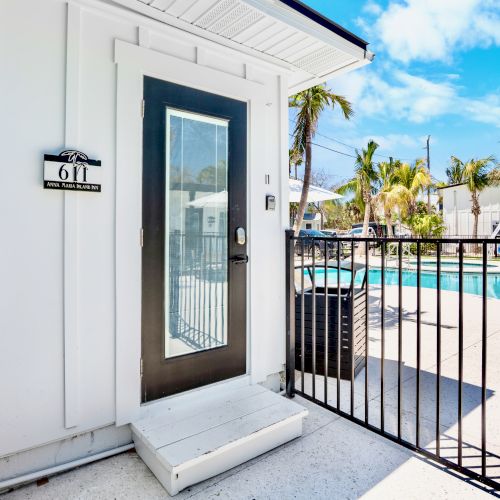 A pool area is visible with a gate, a white building with a door, a fire extinguisher, and palm trees under a blue sky.