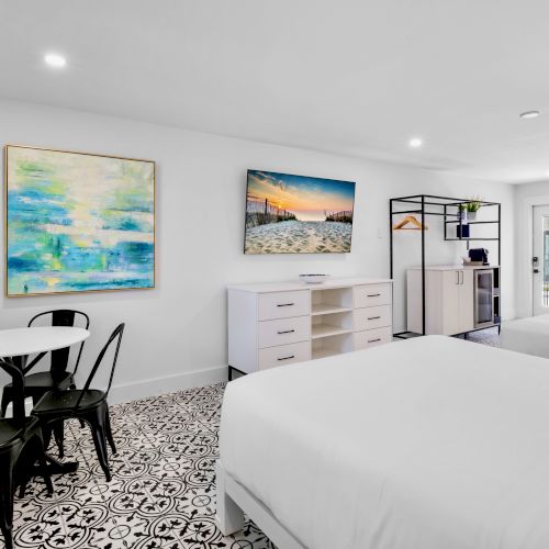 A bright room with two beds, patterned floor, wall art, TV, small dining table, and storage rack.