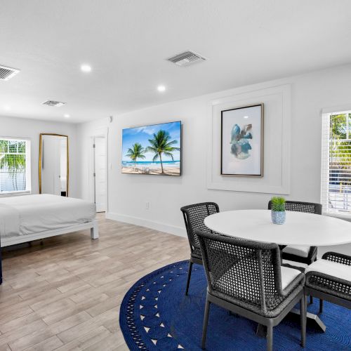 The image shows a bright, modern room with a bed, round table and chairs, beach artwork, and a blue rug, creating a cozy atmosphere.