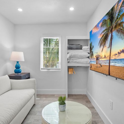 A cozy room with a white couch, a beach scene on the TV, a side table with a lamp, a wall art piece, and a small window with folded towels.