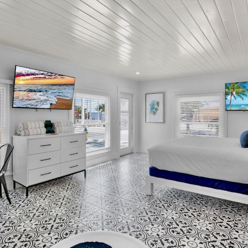 A bright bedroom with patterned floor tiles, a bed, dresser, two wall-mounted TVs, and a small table with a chair by the window.
