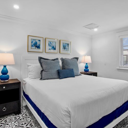 The image shows a modern bedroom with a white bed, blue accents, two lamps, patterned floor, and three artworks on the wall.