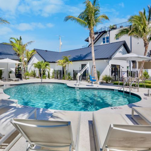 The image shows a serene pool area with lounge chairs, palm trees, umbrellas, and a modern building under a clear blue sky.