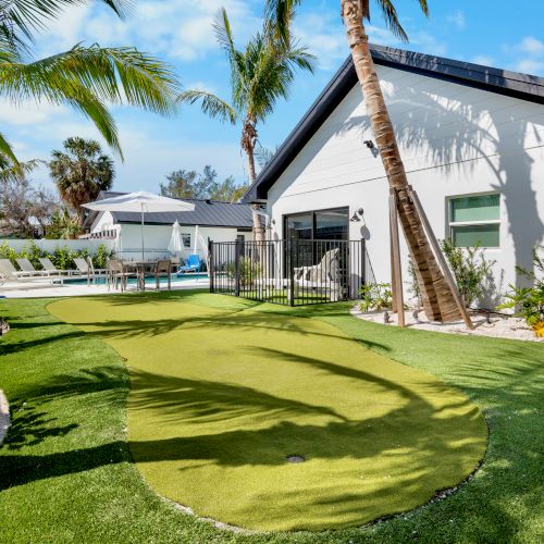 The image shows a backyard with a small putting green, palm trees, a white house, and a fenced-in pool area under a clear blue sky.