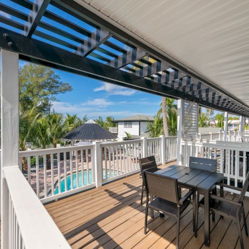 A wooden deck with outdoor seating overlooks a pool and lush greenery, under a blue sky.
