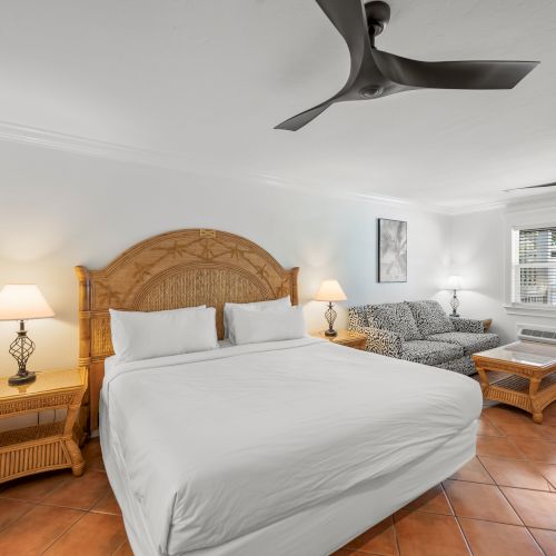 A bright hotel room features a large bed, wicker furniture, two lamps, and ceiling fans, with artwork and a window above a sofa.