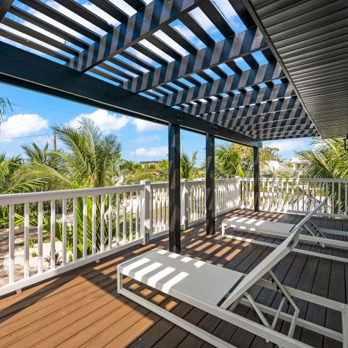 A sunlit patio features two white lounge chairs under a pergola, with palm trees and a blue sky in the background.