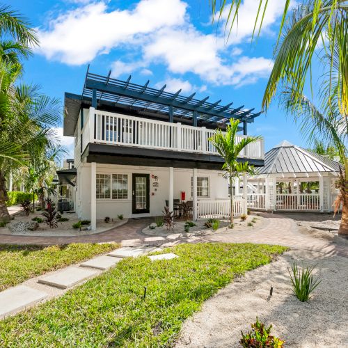 A two-story house with a pergola, surrounded by tropical plants and a stone path in a landscaped garden under a bright blue sky.