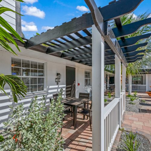 A patio with a pergola, outdoor seating, and plants on a sunny day. There are orange lounge chairs on a tiled surface.