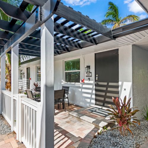 The image shows a modern patio with a pergola, outdoor seating, plants, and a pathway, adjacent to a white building with a black door.