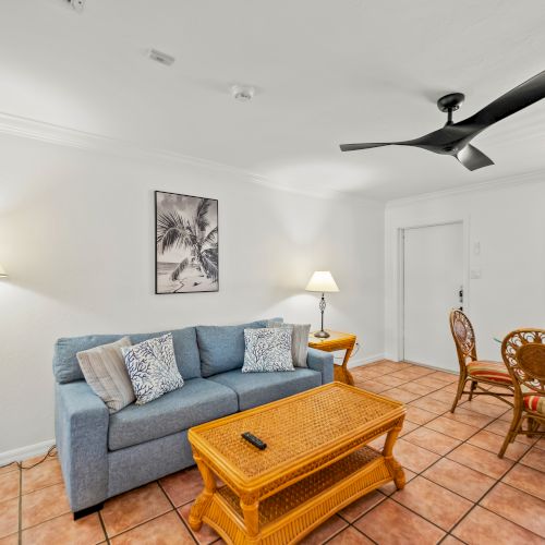 A cozy living room with a blue sofa, wicker coffee table, dining area, and modern ceiling fan, lit by lamps and natural light from a window.
