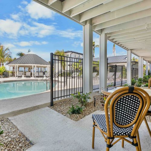 An outdoor pool area with a gated fence, lounge chairs, and a covered patio with wicker seating, sunny sky, and neatly landscaped paths.