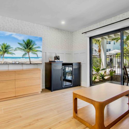 A bright living room with a ceiling fan, light wood floors, a wall-mounted beach photo, a wooden dresser, a black cabinet with a mini-fridge, and a sliding glass door opening to an outdoor patio.