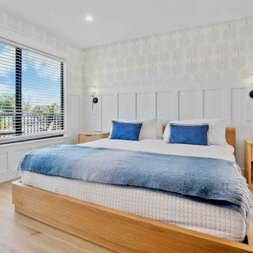 A bright, airy bedroom with a low wooden bed, pale walls, and large windows letting in natural light, creating a calm, minimalist vibe.