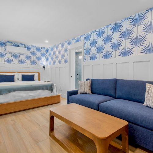 A bright hotel room with a wooden bed, blue upholstered sofa, wooden coffee table, and tropical blue leaf-pattern wallpaper, plus white wainscoting.