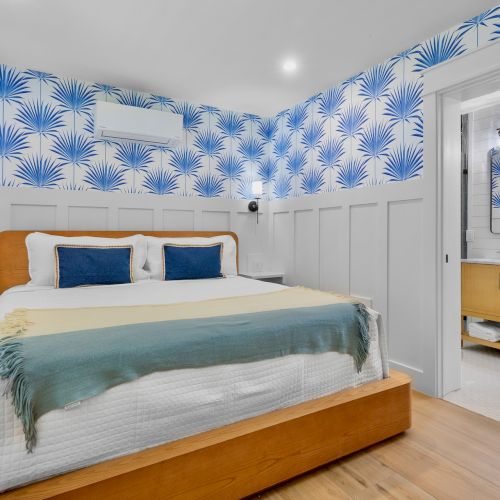 A bright bedroom with a wooden bed, blue palm-patterned wallpaper, light wood floor, white wainscoting, and an adjoining bathroom.