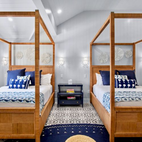 The image shows a bedroom with two canopy beds, blue and white bedding, a central nightstand, and patterned floors in a bright, airy space.