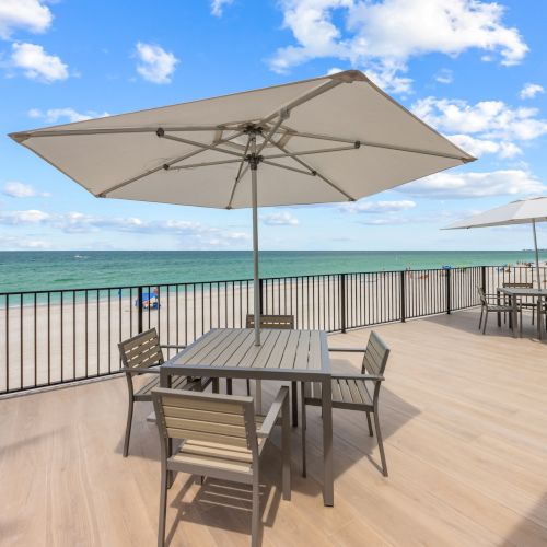 A seaside deck with tables and chairs under umbrellas overlooks a sandy beach and the ocean, with a partly cloudy sky above.