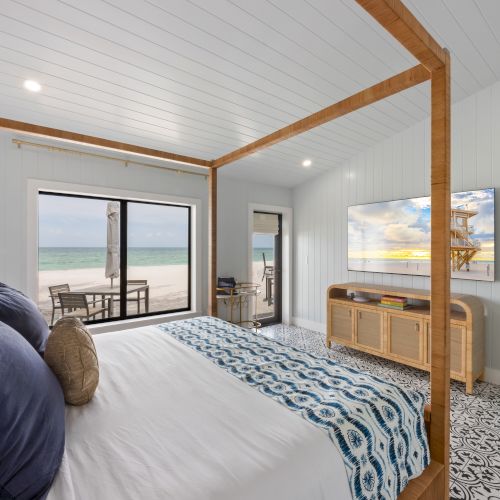 A cozy bedroom with a canopy bed, ocean view window, patterned floor, and a TV. Blue accents complement the serene atmosphere.