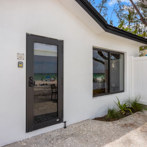 A small white building with a glass door and window, reflecting a beach scene, and a small outdoor area with a table.
