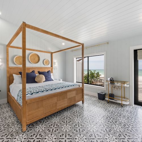 The image shows a bedroom with a four-poster bed, patterned floor tiles, and a view of the beach through a glass door and window.