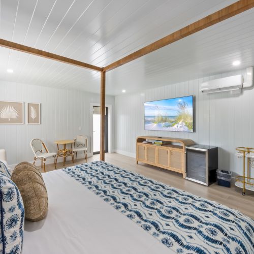 A modern bedroom features a four-poster bed, patterned bedding, wall art, TV, and a seating area, all in a bright, airy space.