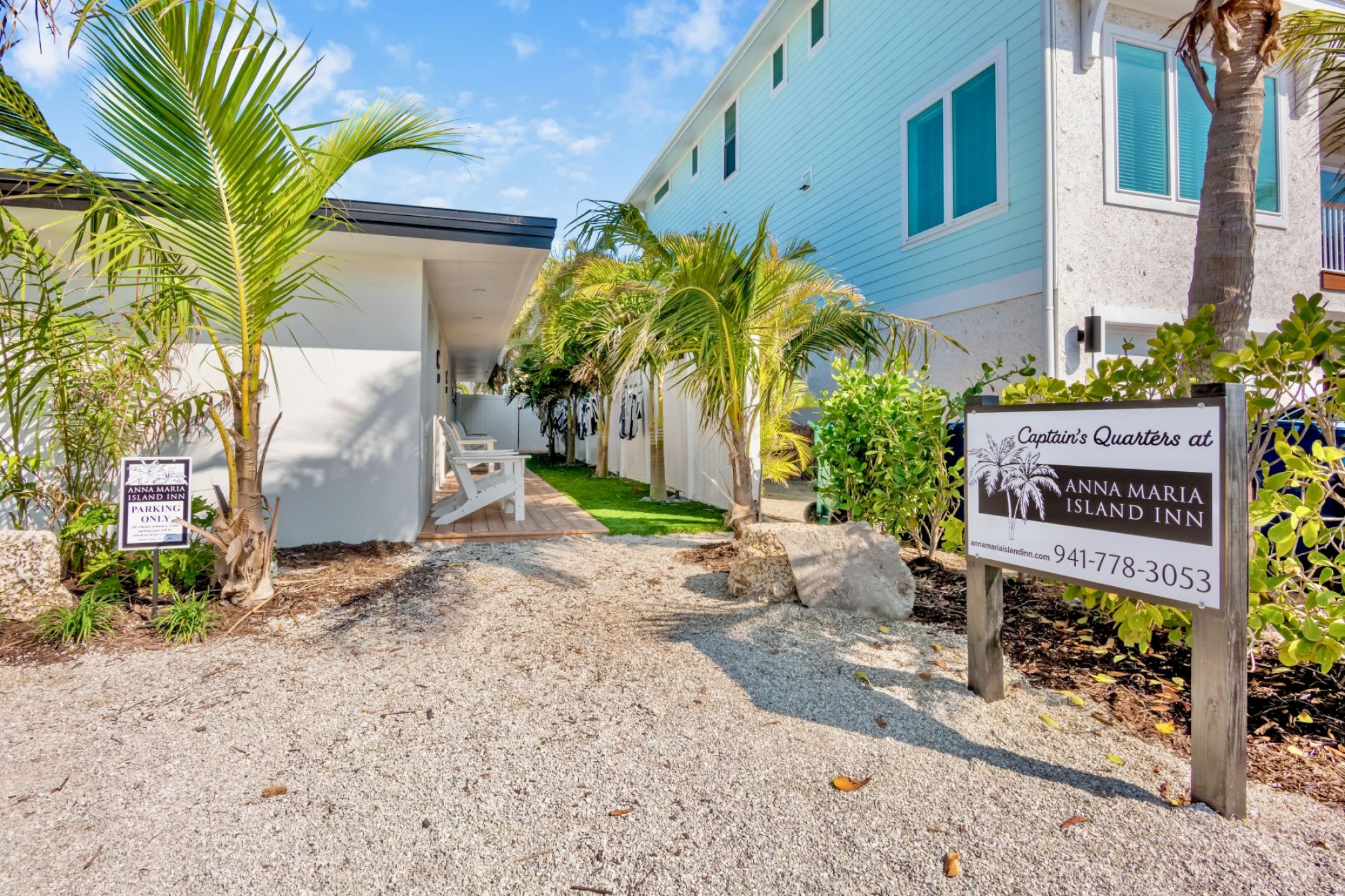 The image shows a walkway with palm trees, leading to a building at Anna Maria Island Inn, with a sign displaying contact information.