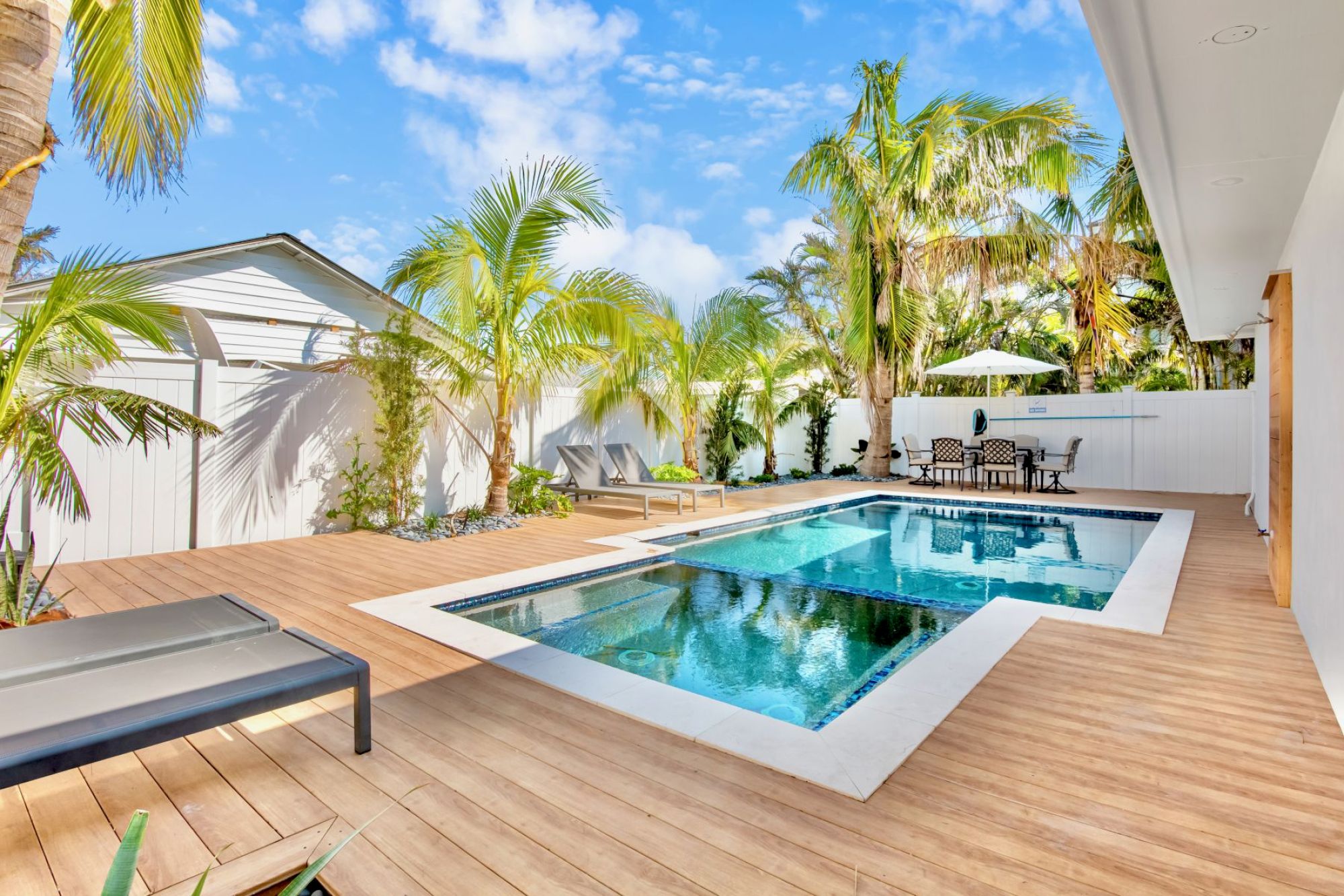 A modern backyard with a swimming pool, wooden deck, lounge chairs, and palm trees under a sunny blue sky.