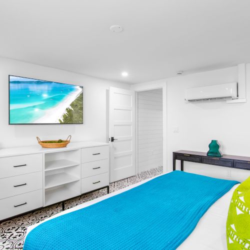 A bright bedroom features a bed with blue and green linens, a dresser with a basket, a wall-mounted TV, and a split air conditioning unit.