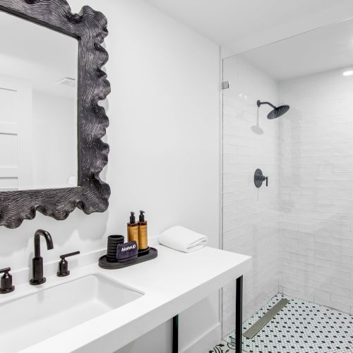 A modern bathroom features a white countertop with a decorative mirror, black fixtures, toiletries, towels, and a glass-enclosed shower.