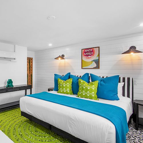 A modern bedroom with a large bed featuring blue and green accents, wall art, a basket of moss on a white counter, and bright lighting.