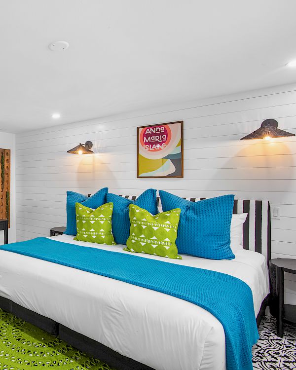 A modern bedroom with a large bed featuring blue and green accents, wall art, a basket of moss on a white counter, and bright lighting.