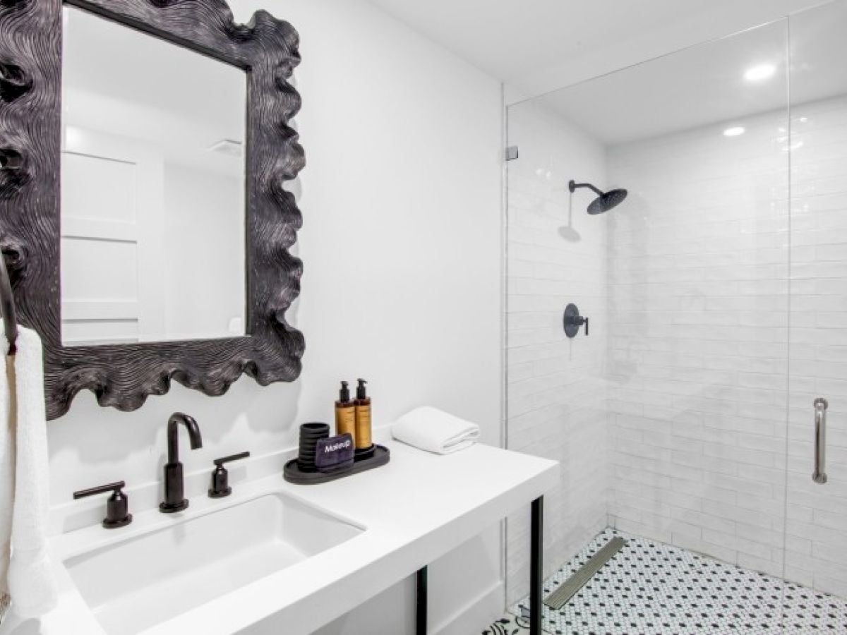 The image shows a modern bathroom with a frameless glass shower, ornate mirror, and black fixtures on a white sink countertop.