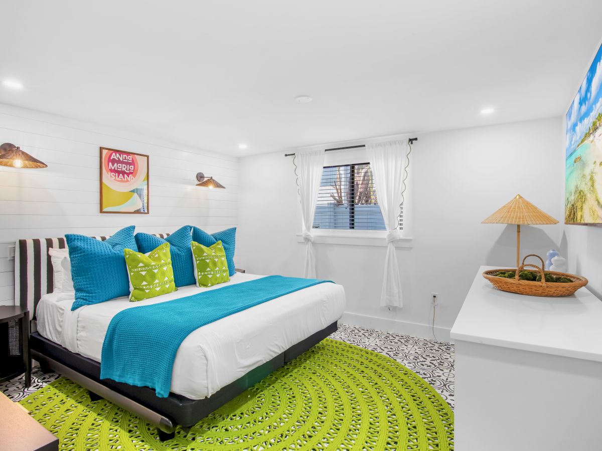 A bright bedroom with a bed, blue and green bedding, wall art, a circular green rug, and a TV. A window provides natural light.