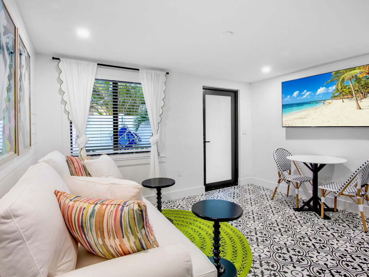 A cozy room features a white sofa with colorful cushions, a decorative rug, a small dining area, and beach-themed wall art, creating a relaxing vibe.