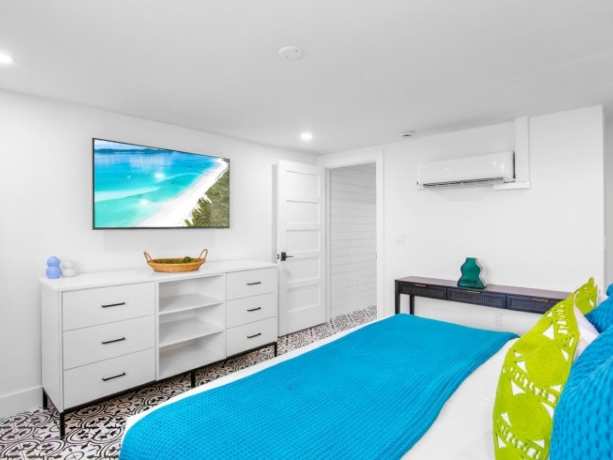 A bright bedroom with a TV, blue bedding, white furniture, and a beach image on the screen.