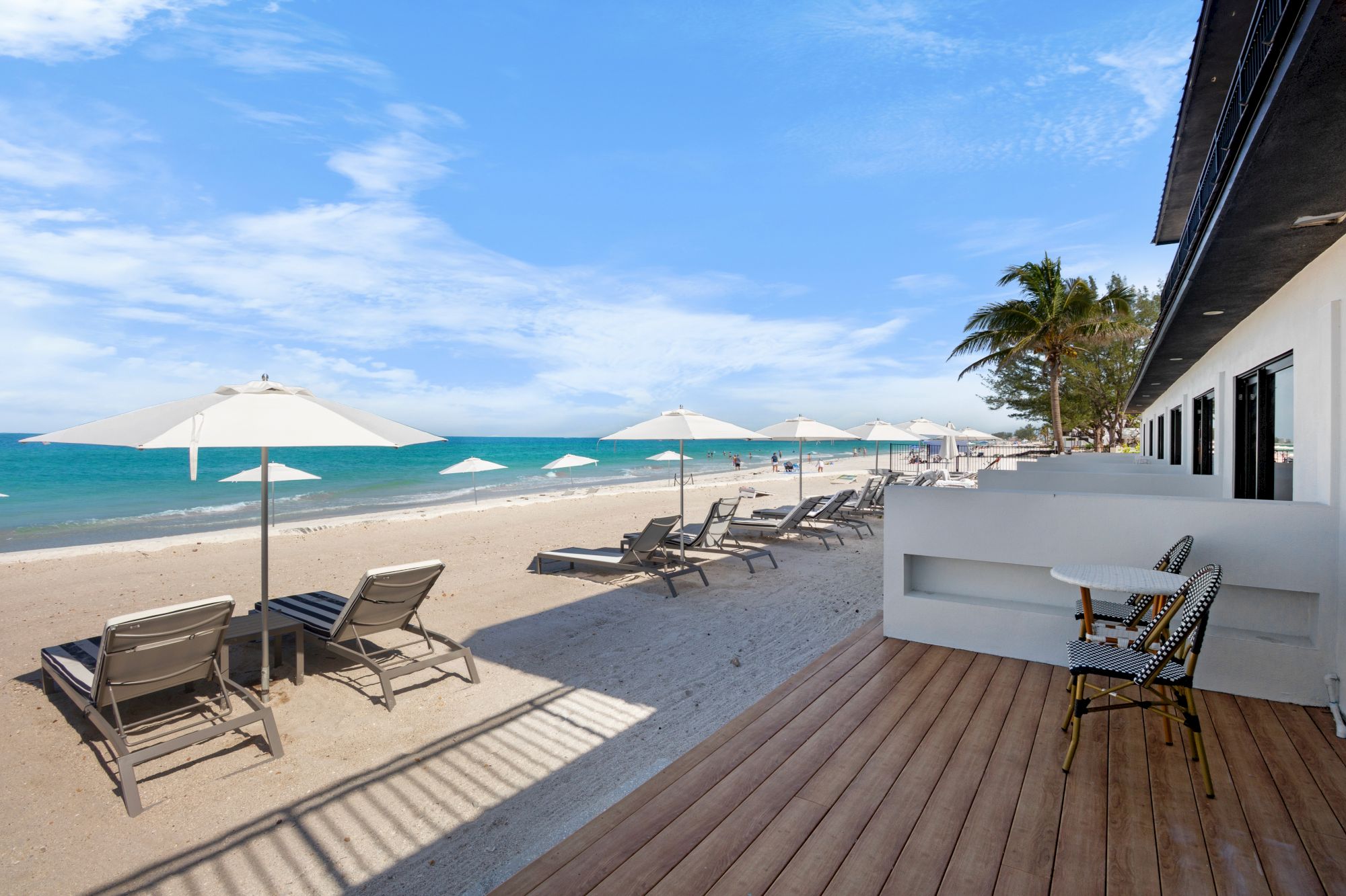 A beach scene with lounge chairs, umbrellas, and a wooden deck next to a building overlooking the ocean under a partly cloudy sky.