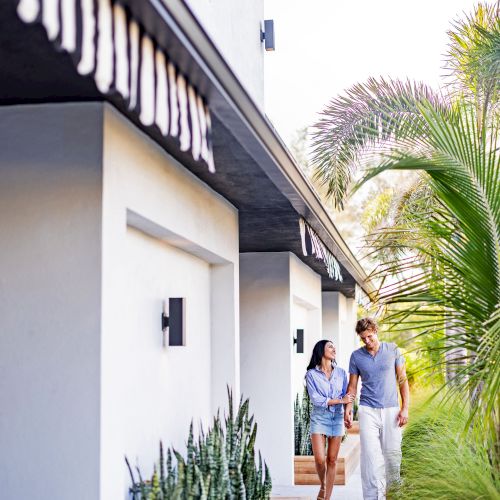 A couple is walking outside a modern house with plants and trees along the path under a clear sky.