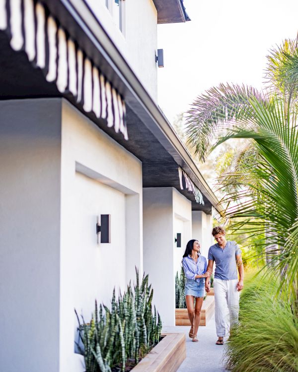 A couple is walking outside a modern house with plants and trees along the path under a clear sky.