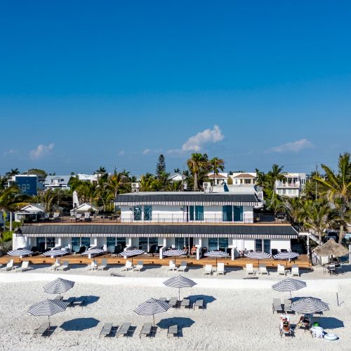 A beachfront scene with a building, sun loungers, and umbrellas on the sand, surrounded by palm trees and blue sky.