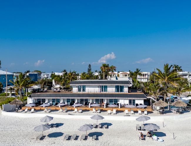 A beachfront scene with a building, sun loungers, and umbrellas on the sand, surrounded by palm trees and blue sky.