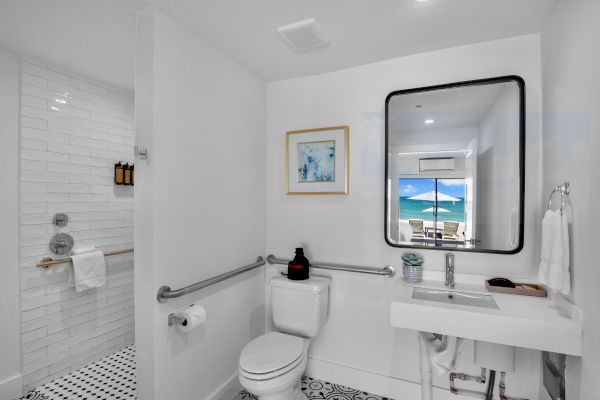 A bright bathroom with a toilet, sink, shower, and large mirror. The window shows a scenic beach view in the background.