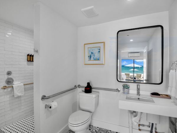 A bright bathroom with a toilet, sink, shower, and large mirror. The window shows a scenic beach view in the background.