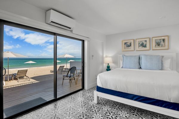 A beach-view bedroom with a bed, decorative pillows, art on the wall, and a glass door opening to a deck with chairs and umbrellas overlooking the sea.