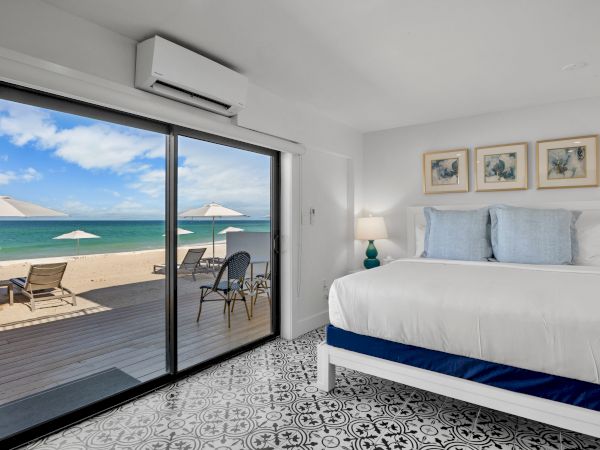 A beach-view bedroom with a bed, decorative pillows, art on the wall, and a glass door opening to a deck with chairs and umbrellas overlooking the sea.